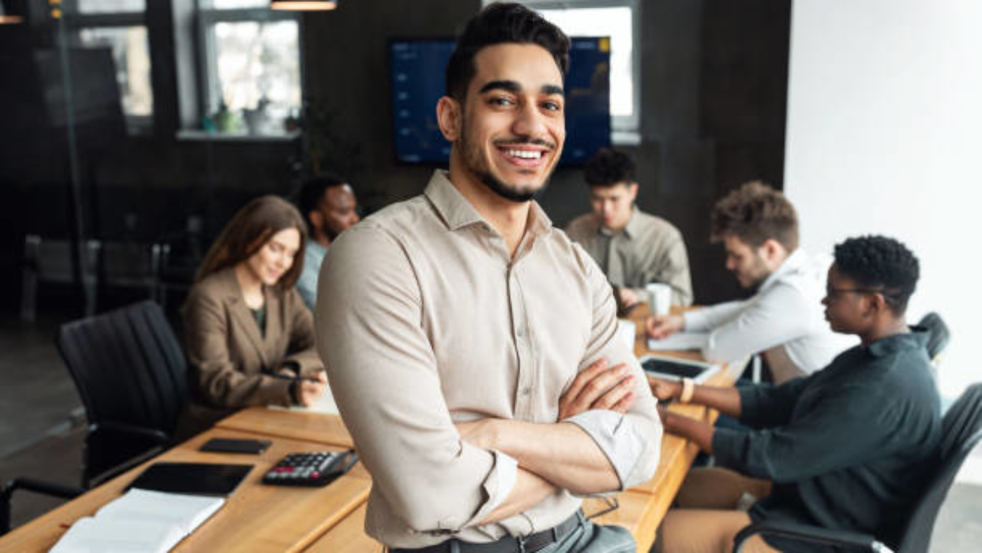 Successful Person. Portrait of confident smiling bearded businessman sitting leaning on desk in office, posing with folded arms and looking at camera, colleagues working in blurred background